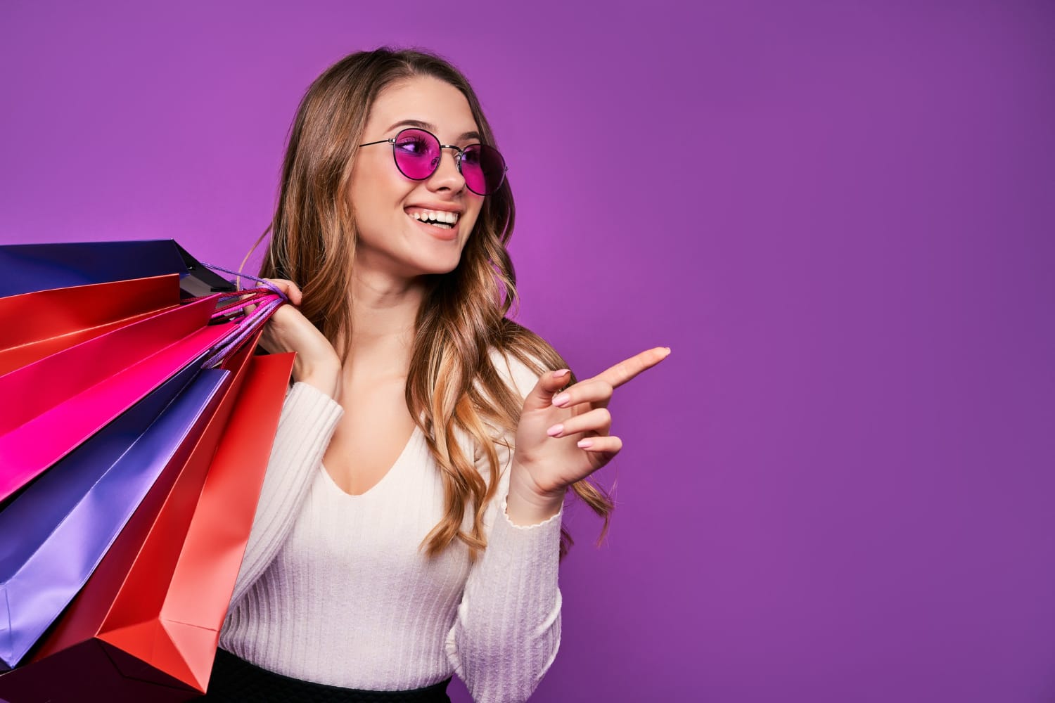 Mulher sorridente com óculos de sol rosa segurando sacolas de compras coloridas, em frente a um fundo roxo, simbolizando uma grande promoção na loja Concept Áurea.