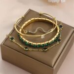 Pulseiras douradas Concept Áurea com pedras verdes e detalhes em strass sobre tecido sofisticado.